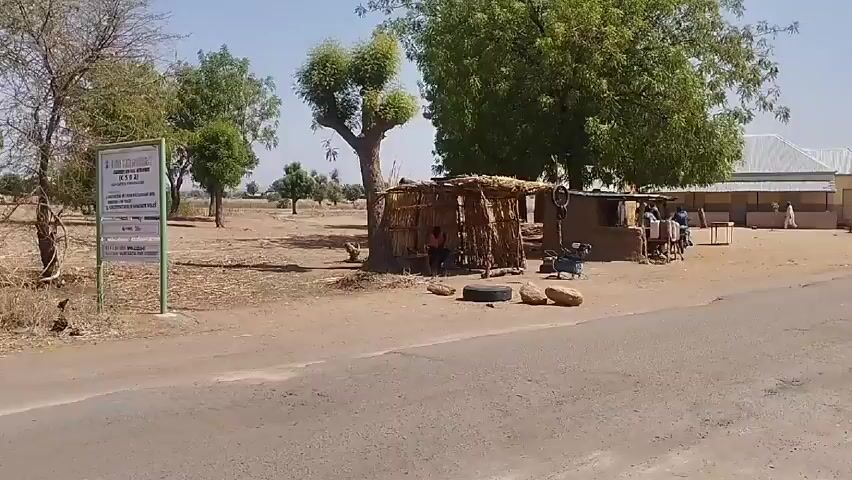 The video presents a daytime scene in what appears to be a rural setting. The camera, in motion, captures the landscape from the perspective of a vehicle traveling along a paved road. The environment features sandy, dirt terrain dotted with trees. Structures are visible, including simple, makeshift huts made of natural materials like straw and wood, as well as a larger, more substantial building with a galvanized metal roof. People are present, some gathered near the structures and others riding motorbikes. The overall impression is one of a small, possibly impoverished village community.