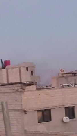 The video shows the rooftops of several buildings. The buildings are made of beige bricks. On the rooftops, there are satellite dishes, water tanks, and people. The sky is overcast. The camera is shaky.