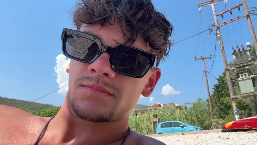 The video opens with a close-up shot of a young man with curly hair and sunglasses, looking directly at the camera. He is shirtless and on a beach. In the background, there are trees, a utility pole with electrical equipment, and cars. The camera then pans to show the man lying on a beach towel next to a young woman who is reading a book. She is wearing a bikini and sunglasses. The man then turns back to the camera and makes a rock-and-roll hand gesture. The video continues with more shots of the man and woman on the beach, with the man occasionally looking at the camera. The environment appears to be a sunny day on a rocky beach.