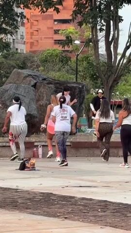 A group of people are gathered in an outdoor area, possibly a park. A woman in a white outfit is standing on a raised platform and appears to be leading an exercise class or dance session. She is gesturing with her hands and seems to be speaking or singing into a microphone. Several other women, dressed in athletic wear, are dancing or following along with her movements. One person in the background is holding a camera or phone, possibly recording the event. There are trees and buildings visible in the background. The ground appears to be paved. There are a few water bottles on the ground near the platform.