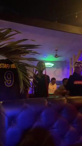 The video shows a dimly lit indoor space that appears to be a bar or club. There are several people present, some standing and some seated. In the foreground, a black leather sofa is visible. To the left, a large green palm frond partially obscures the view. In the background, there are neon signs, including one that says "JAMESON". A woman in a red patterned dress with a long braid is dancing or moving in the center of the frame, smiling and looking towards the camera. To her right, a person wearing a black hoodie with "Supreme" written on it is visible. Other individuals are blurred or partially visible in the background. Bottles and red cups are on a table in the foreground. The lighting is a mix of ambient light and colored spotlights, creating a vibrant atmosphere.
