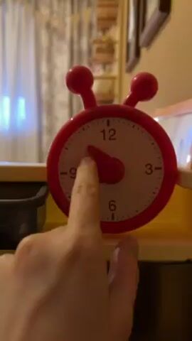A hand is adjusting the short hand on a red plastic clock with a white face. The clock features two red balls on top and numbers marking the hours. The clock is resting on a yellow surface. In the background, a curtain, a shelf with various items, and a picture frame can be seen.