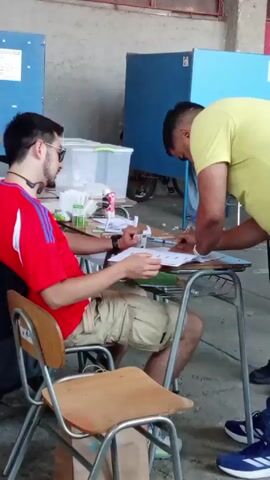A man in a yellow shirt is standing at a school desk and watching a man in a red shirt sit at the desk. The man in the red shirt is holding a document and appears to be showing it to the other man who is pointing at something on the document. The man in red is wearing sunglasses and has a headset around his neck. The man in yellow has a short haircut.  There are a few other people visible in the background, as well as some blue voting booths. The setting appears to be an indoor space, possibly a school or community center, with grey walls. The scene likely depicts a voting process, with the man in yellow possibly assisting the man in red.