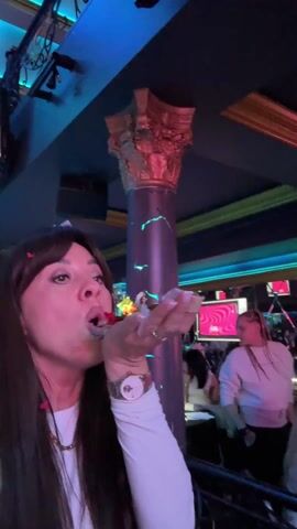 A woman with long dark hair and bangs wearing a white long-sleeved shirt, a watch, and a necklace, blows something, potentially confetti or glitter, off her hand in a nightclub setting. The lighting changes colors, primarily between green and pink. In the background, other people are visible, and there is a bar area and decorative architectural details such as a gold-colored pillar and wrought iron railing. A screen displays colorful graphics.