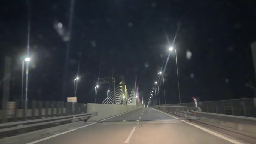 The video shows the perspective from inside a car driving at night on a bridge, with the lights on the bridge illuminating the structure and the road. The bridge is supported by a large pylon with many supporting cables. The pylon is lit with colored lights. Street lights line the bridge on either side of the road. There is no other traffic visible. The windshield of the car has some water droplets on it.