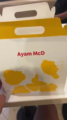 A person opens a white and yellow "Ayam McD" box and reveals chicken bones. The box sits on a table. Some trash is in the background.