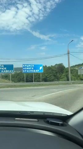 The video begins with a view from inside a vehicle, looking out at a road. Two blue road signs are visible. The left sign indicates "ANAPA TEMRYUK PORT KAVKAZ" with an arrow pointing left. The right sign indicates "KRYMSK ABINSK KRASNODAR" with an arrow pointing right. The signs are mounted on poles above the road. Beyond the signs, there is a landscape with greenery and buildings in the distance. The sky is blue with white clouds.

The video then transitions to show a wider view of the environment. Two tall lampposts with wires are prominent. A street corner is visible, with a few more signs and what appears to be a small building or structure. The landscape continues to be visible in the background, with rolling hills and vegetation. The view is again from within a vehicle.