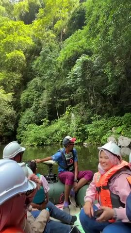 A group of people are on a boat in a body of water, surrounded by lush green trees and vegetation. The boat appears to be a raft or a small inflatable boat. Some of the people are wearing life vests and helmets. One person in the back is steering the boat with a motor. The person in the foreground on the right is smiling and holding a camera or phone. The environment appears to be a natural setting, possibly a river or a lake within a jungle or forest. The water is calm and reflects the surrounding greenery. The overall mood seems to be one of enjoyment and exploration.