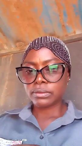 A person with dark skin, wearing a gray collared shirt and black framed glasses, looks directly at the camera. The person is wearing a patterned headscarf. Behind the person is a weathered wall and roof. A phone watermark is visible in the bottom left corner. The person slightly moves their head from side to side.