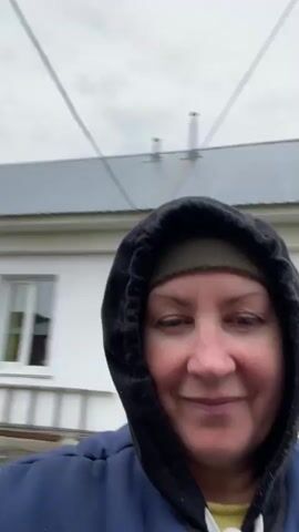 A person wearing a blue jacket with a black hood over a green head covering looks at the camera and smiles slightly. The person is outside, in what appears to be a rural or suburban environment. Various structures, including a wooden building, a fence, and other outbuildings, are visible in the background. The sky is overcast. The video appears to be taken with a hand-held camera, as there is slight movement in the frame.
