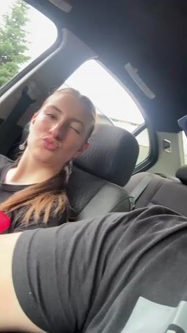 The video shows a young woman with brown hair in a ponytail, wearing a black t-shirt with a red design on it, and black shorts. She is sitting in the back seat of a car. The car's interior is visible, including the headrest of the seat in front of her and the door frame. The window is open, and trees and a building can be seen outside. The woman is looking at the camera and making various facial expressions and gestures, including talking, flexing her arm, and smiling. There is another person's leg in the foreground, wearing black shorts. The video appears to be a selfie taken from the perspective of the person in the front seat.