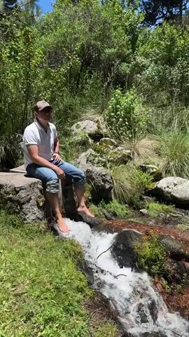A man is sitting on a stone edge with his feet in a small stream. He is wearing a white polo shirt, blue jeans, and a camouflage baseball cap. The stream is surrounded by lush green grass and trees. The water in the stream is clear and flowing over rocks. Sunlight filters through the trees, creating dappled shadows on the ground. The man appears relaxed as he dips his feet into the cool water. The scene is peaceful and natural, evoking a sense of tranquility.