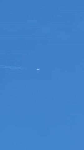 A small white airplane is visible in the distance against a bright blue sky. The airplane is leaving a long white contrail behind it, which stretches across the frame. The contrail is fading and becoming less distinct towards the right side of the image. The sky is clear except for a few wispy white clouds scattered throughout. In the last frame, a dark wire crosses the top of the image horizontally.