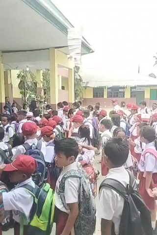 A group of young children, dressed in school uniforms and wearing backpacks, are gathered outdoors in what appears to be a schoolyard. Many of the children are wearing red hats. They are standing in rows, facing towards a building. There are adults present, some of whom appear to be teachers or staff. The building has yellow walls and white pillars. The sky is visible and appears to be overcast. There are some decorations hanging from the roofline of the building, including what looks like paper streamers. The children are mostly looking forward, with some interacting with each other. The overall impression is one of a school assembly or a gathering before or after classes.