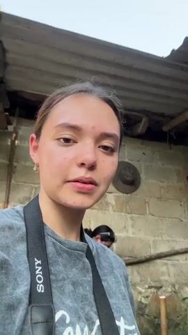 The video shows a young woman recording a vlog-style video outdoors. She's standing with a man in an olive-green t-shirt with the word "ARQUITECTXS DESCALZXS" on it, and he poses for the camera as she shows her face and smiles. She is wearing a grey t-shirt and a camera strap that reads “SONY” around her neck. Behind them, other people are milling around a small construction site in a lush, wooded area. Some are carrying buckets and tools, while others are walking up and down a set of steps. The construction site appears to be adjacent to a building with brick walls and a tin roof. The woman is looking directly at the camera and talking, while occasionally panning the camera to show the surrounding environment and the people working in it.