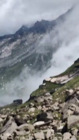 The video shows a mountainous landscape with fog rolling in. The foreground is filled with rocks and sparse green vegetation. The middle ground features a steep, rocky slope dotted with patches of green. The background is dominated by a large, rugged mountain peak partially obscured by clouds and fog. The overall impression is one of a wild and dramatic natural environment.