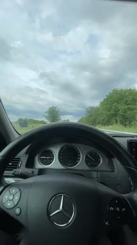 The video shows the interior of a Mercedes-Benz car, viewed from the driver's perspective. The steering wheel with the Mercedes-Benz logo is prominent in the foreground. The dashboard displays gauges, including a speedometer. The car is on a road, with trees and greenery lining the sides. The sky is cloudy. The car appears to be in motion.