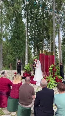 A wedding ceremony is taking place outdoors in a forest setting. Tall trees surround the area. A wedding arch decorated with red and green floral arrangements stands behind the bride and groom. The bride wears a white wedding dress and a veil, and the groom wears a dark suit. A wedding officiant stands to the left of the couple, holding a microphone and speaking. Guests are seated on green stools arranged in a semi-circle facing the ceremony. Several guests are clapping. The ground is paved with stones. String lights are visible overhead.