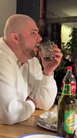 A man with a shaved head and a beard is seated at a wooden table. He's wearing a white hoodie. He holds a short, faceted glass containing a dark liquid. A bottle of beer labeled "ХАДЫЖЕ", a bottle of Coca-Cola, and some crumpled napkins sit on the table. The man looks at the glass, then takes a drink. The background is dimly lit, with a glimpse of a doorway and some plants. Loud music is playing.