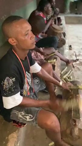 The video shows a group of men sitting in a row on a concrete ledge. The man in the foreground is wearing a black and white t-shirt, denim shorts and a red lanyard. He is using a machete to chop pieces of sugar cane on a wooden block.  Behind him, the other men are playing drums, smiling and cutting the sugar cane as well. The setting appears to be an open-air structure with concrete floors and pillars. The scene conveys a sense of community and traditional music-making.