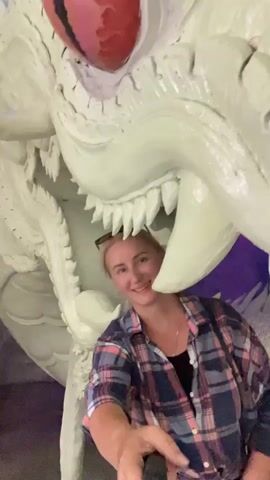 A medium-shot video shows a woman with blonde hair and sunglasses on top of her head smiling while holding a selfie stick. She is wearing a pink, blue, and white plaid shirt over a black tank top. She is positioned in front of a large, white dragon sculpture with red eyes. The dragon appears to be lunging forward, with its mouth open, revealing sharp teeth. The background is dark and appears to be a cave or an indoor space with some purple lighting. The woman is looking at the camera and appears to be taking a selfie video.