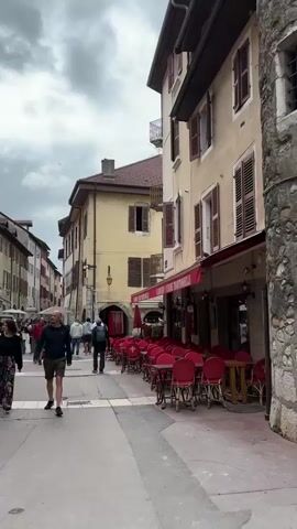 A street scene in a European town with people walking and outdoor seating for a cafe. The buildings are old with colorful facades and shutters. The sky is cloudy. A man in a blue shirt and khaki shorts walks toward the camera. A group of people walk away from the camera. The cafe has numerous red chairs and tables set up.
