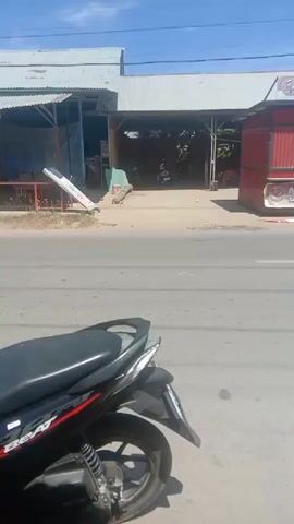 A light-colored scooter carrying two individuals, one driving and one passenger, moves from left to right across the frame, passing a red food stall labeled "AYAM PENYET" and a building with a metal roof. The scene takes place outdoors during daylight with a clear blue sky. In the foreground, the rear of a black and red motorcycle is visible.