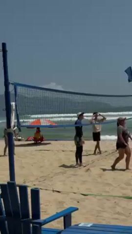A group of six people are playing beach volleyball on a sandy beach with the ocean in the background under a clear sky. The camera is positioned behind a set of blue wooden chairs, partially framing the scene. The volleyball net is blue and white, stretching across the middle of the beach. The ball is mid-air, a yellow and black object arcing over the net. The players are dressed in various beach attire, shorts, t-shirts, and some wearing wetsuits. They are actively engaged in the game, moving and positioning themselves for the ball. The waves of the ocean gently roll onto the shore in the background. A red beach umbrella can be seen further down the beach.