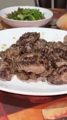 A close-up shot shows a white plate piled with small, grilled meatballs. The meatballs are browned and have visible grill marks. Behind the plate, there is a white bowl of green salad with some red vegetables. To the right of the salad, there is a round, crusty bread roll. The background is blurry but appears to be a dining table setting. The focus is on the meatballs.