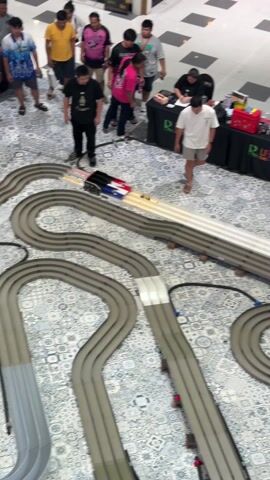 The video shows a slot car race in progress on a gray plastic racetrack, with onlookers gathered around. The track includes straightaways and looping curves, with red and white accent markings.  Several individuals stand behind a registration table displaying a “R Lifestyle” logo.  A person in a white shirt walks along the starting straightaway, while another crouches to retrieve a vehicle near the track.  The patterned floor tiles contrast with the various clothing styles of the crowd.