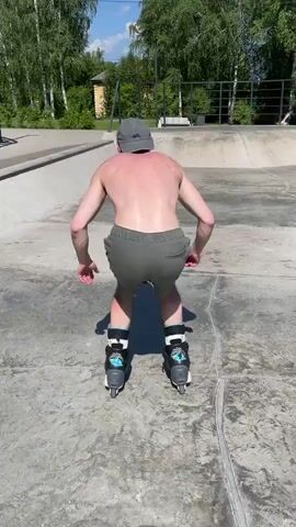 This video shows a person rollerblading at a skatepark on a sunny day. The person, wearing shorts and a baseball cap, is shirtless and appears to be performing tricks on the rollerblades. They are seen grinding on a metal rail in the park, with trees and a building visible in the background. The overall scene suggests a casual outdoor activity in a recreational setting. The time of day is clearly daytime, indicated by the bright sunlight and shadows. The weather appears to be warm and pleasant, suitable for outdoor sports. The location, identified as Divnogorsk, Russia, provides specific geographical context to the scene. No text is visible in the video.