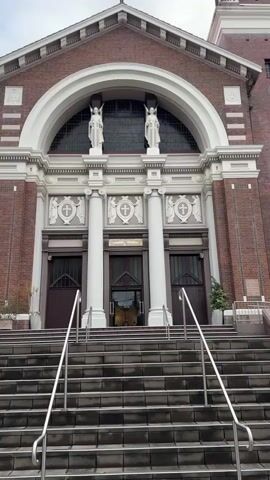 This media file shows the exterior of a brick church with a grand arched entrance. The church features white columns, statues of angels above the entrance, and a large arched window. In front of the church is a long set of stairs with metal railings leading up to the doors. To the right of the church, a bell tower is visible, and a banner displaying "WELCOME" is attached to a lamppost. Two potted plants, one on each side of the entrance, add a touch of greenery. The sky is overcast, suggesting a cloudy day. The setting appears to be an urban or suburban area, and the architecture suggests a place of worship. There are no people visible in the frame, and the overall mood is serene and inviting.