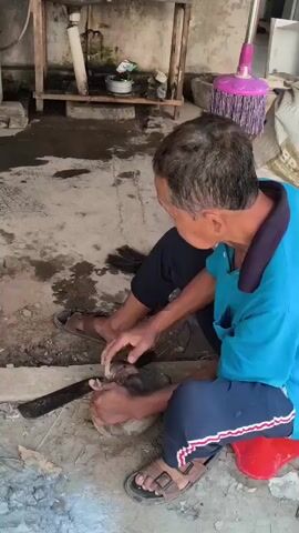 An elderly man wearing a blue shirt and dark pants is sitting on the ground in an outdoor area. He is holding a piece of wood in his left hand and appears to be using a tool to shape or clean it with his right hand. In front of him is a rough, round stone, likely a grinding stone or mortar. To his left, there is a large, dark, curved blade, possibly a machete or sickle. Behind him, a pink mop stands upright against a wall. The ground is dirt and concrete, with some debris and water stains. A wooden shelf with items on it is visible against a wall in the background.