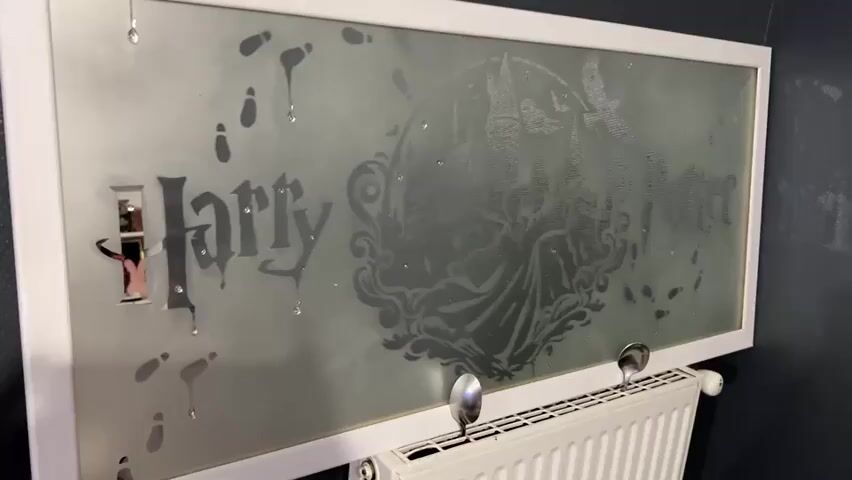 The video shows a close-up shot of a frosted glass panel with a white frame. The panel is decorated with a Harry Potter theme, featuring footprints, the Hogwarts castle, and the name "Harry Potter" in stylized lettering. There are also small, sparkly embellishments scattered across the glass.

On the left side of the panel, the word "Harry" is visible in a bold, dark font. Some of the letters appear to be made of mirrored material, reflecting the surroundings. There are also several teardrop-shaped marks on the glass, some with small droplets of liquid clinging to them.

To the right of "Harry", a circular design depicts Hogwarts castle, with a figure in a cape and a bird flying above. The name "Potter" is written in a similar dark, stylized font to the right of the castle.

Below the glass panel, a white radiator is visible, with two metal spoons attached to the top of it, facing upwards. The background is a dark, textured wall. The lighting in the scene is dim, creating a slightly mysterious atmosphere.