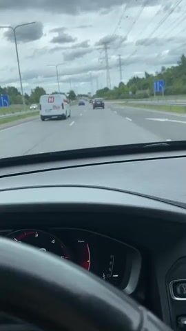 The video shows a view from inside a car driving on a highway. The dashboard displays a speedometer reading of 77, then 76, and finally 75. The road has multiple lanes and is lined with trees and power lines. Several other vehicles are visible on the road, including a white van with a red logo and a few cars. The sky is cloudy.