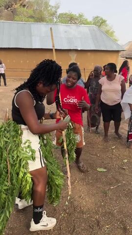 The video shows a woman with curly hair, wearing a black tank top and white shorts, fastening a bundle of leafy branches to a long, thin pole. She is smiling and surrounded by a small crowd of people in an outdoor area with a dirt ground. Some of the people are also holding leafy branches and look to be participating in a ceremony or performance. In the background, there is a building with a corrugated metal roof and a small hill or rocky outcrop. Other people are watching and filming. The woman with the leafy branches then looks directly at the camera and smiles.