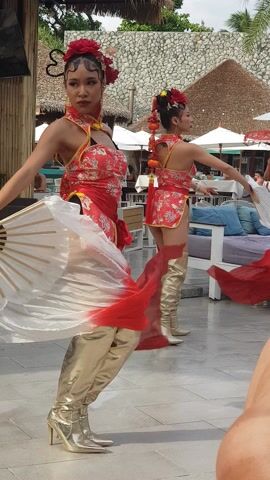 Two women dressed in red Chinese-style dresses and gold thigh-high stiletto boots dance with red-and-white fabric fans on a patio. The patio is near palm trees and a thatch-roofed building, and there is patio furniture visible behind the women. The women’s hair is styled in a bun with red floral accessories.