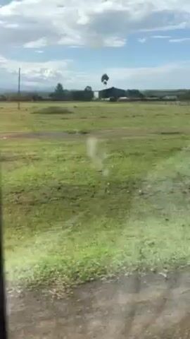 From a moving vehicle, likely a bus, a flat grassy landscape is visible through the window. Scattered buildings and trees dot the horizon under a partially cloudy sky. The landscape appears rural and possibly African, based on the style of buildings and vegetation. The window is somewhat blurry. A significant amount of wind noise is audible in the recording.
