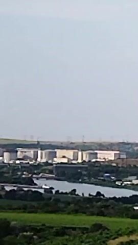 A slow-panning shot shows an industrial area with large, light-colored cylindrical tanks situated next to a body of water. Beyond the industrial area, a rolling landscape with green vegetation, fields, and trees is visible. In the distance, several wind turbines are scattered across the horizon against a pale blue sky. The camera moves from left to right, capturing the vast expanse of the scenery.