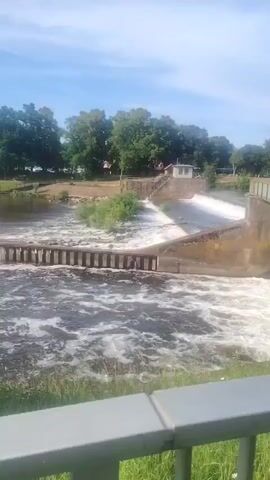 The video shows a wide shot of a dam with water flowing over it. The dam appears to be made of concrete and has a bridge crossing over it. There are trees and buildings in the background. The water is choppy and white with foam, indicating a strong current. The sky is blue with some clouds. The video is shot from a slightly elevated position, looking down at the dam and the water. There are no people visible in the video. The overall impression is of a powerful natural force being controlled by man-made structures.