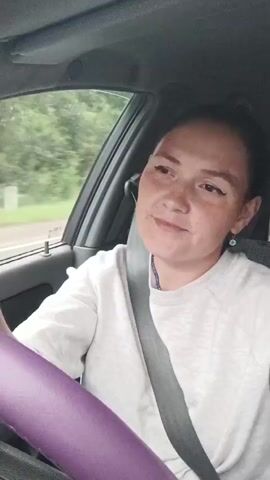 A woman is driving a car and looking at the camera. She has dark hair pulled back in a bun and is wearing a white sweatshirt and a seatbelt. Her left hand is on the steering wheel, which is covered in a purple material. She appears to be smiling faintly. The background shows trees and greenery, indicating she is on a road surrounded by nature. The lighting suggests it is daytime.