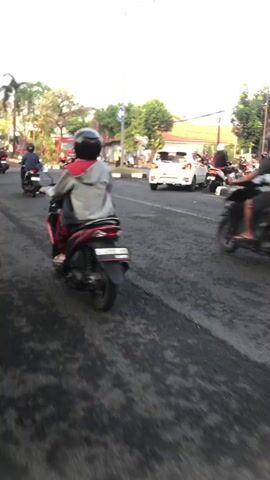 The video shows a street scene with several motorcycles and a white car. The perspective is from a moving motorcycle, looking forward. In the foreground, the asphalt road is visible with some water stains. Multiple motorcycles are seen from behind and from the side. One motorcycle in the center foreground has a rider wearing a dark helmet and dark clothing. In the middle ground, a white car is turning. Further in the background, a red truck is visible, along with traffic signals and utility poles. The environment appears to be a city street with buildings and trees on the sides. The lighting suggests it is daytime, possibly late afternoon. There is movement of traffic.