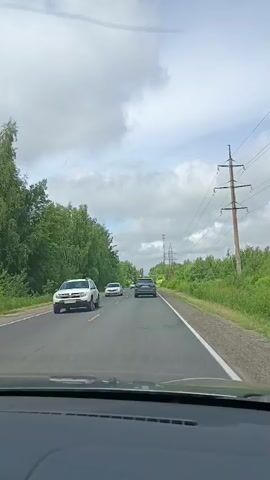 The video is a first-person view from inside a car driving on a two-lane asphalt road. The sky is cloudy with patches of blue. On the left side of the road, there is a dense line of green trees. On the right side, there are also trees and a field, with a power line pylon visible in the distance. Several cars are on the road. A white car is ahead on the left lane, followed by a dark-colored car. Another dark-colored car is ahead on the right lane. A white car with red accents passes the viewer's car on the left lane. Later, a dark SUV passes the viewer's car on the left lane. The road has yellow and white lane markings.