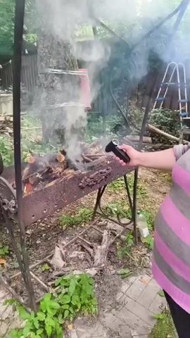 This video shows a person, likely female based on the arm and hand, using a small, black torch to ignite a fire in a metal brazier. The brazier is filled with firewood, some of which is already burning. Flames are growing larger, and smoke is rising. The scene appears to be outdoors, possibly in a backyard or garden area, with greenery and trees visible in the background. A ladder and some sort of large container are also in the background, along with a plastic bottle on the ground. The ground is covered with dirt and some paving stones. The overall impression is that of someone preparing for or actively starting a barbecue or outdoor fire. There is no visible text in the video.