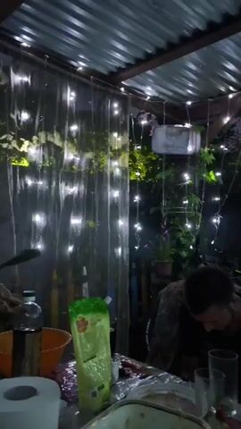 The video begins with a shot of a person sitting at a table outdoors, illuminated by string lights. The person is wearing a dark shirt and appears to be looking down. The table is set with food and drinks, and there are several glasses and plates visible. In the background, there are plants and more string lights. The camera then pans up to show a makeshift shelter with a corrugated metal roof. A swing with a bag hanging from it is visible overhead. The lighting is dim, and the overall atmosphere is relaxed and casual.