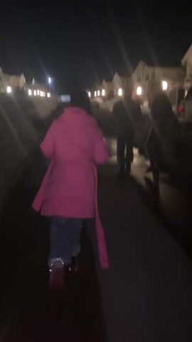 At night, three people walk uphill away from the camera along an icy asphalt road. The person closest to the camera is wearing a bright pink knee-length coat. The other two people, both male, are wearing dark clothing and walk side by side. There are piles of snow on both sides of the road, and on either side, two-story townhouses with small porch lights line the street. At the top of the hill, a streetlight can be seen.