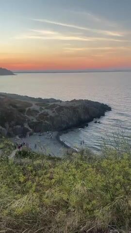 A video taken at dusk shows a coastal landscape with a rocky shore and a small beach. The sky is a gradient of orange and pink, transitioning into blue at the top. The sea is calm, with gentle waves lapping at the shore. There are a few people on the beach, some swimming or wading in the water, and others sitting or walking on the sand. Several small boats are anchored in the water. In the foreground, there are green bushes and dry grass. The camera appears to be positioned on a hill overlooking the scene.