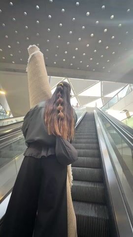A woman in black, carrying a large rolled rug, rides up an escalator. She is wearing a black leather jacket, a black purse, black pants, sunglasses, and gold earrings. She has long brown hair with some light brown highlights, pulled back and held in place with several barrettes. The escalator is surrounded by glass and metal and a concrete ceiling with round light fixtures.