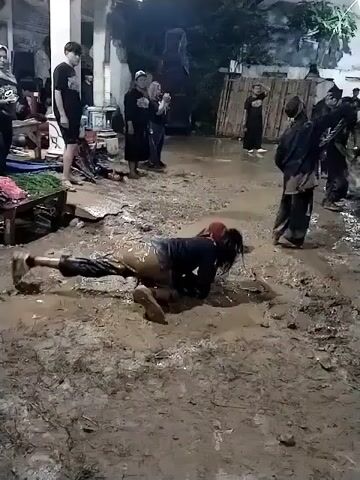 The video captures a nighttime scene in Jenggrik, Indonesia, where a person is performing an activity in a very muddy outdoor area. The central figure, wearing a dark long-sleeved shirt and light-colored pants, is initially seen crawling on their hands and knees through thick mud. They have long dark hair that appears wet and matted.

After crawling, the person stands up and walks away through the mud, carrying a folded red mat or fabric over their left shoulder. The ground is entirely covered in wet, slippery mud, reflecting the artificial lights illuminating the scene.

In the background, several other people, mostly adults, are observing the activity. Some are standing near a white building with pillars, while others are closer to the muddy area. Some observers are wearing black clothing, and some women are wearing headscarves. A blue tarp and a table with what appears to be green leaves or vegetables are visible near the building. The overall atmosphere suggests a communal event or performance taking place in challenging conditions, possibly after heavy rain, or the mud is intentional for the activity. There is no visible text.