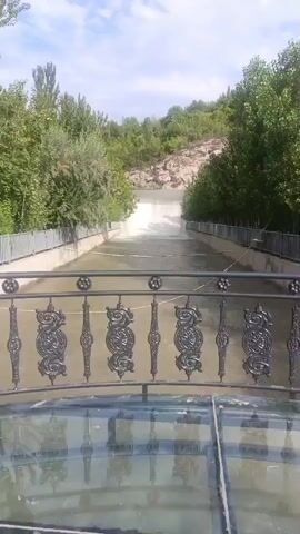 The video shows a body of water flowing through a channel. The channel is lined with concrete walls and metal railings. Trees and vegetation grow on the slopes above the channel. The water appears to be flowing downhill towards a drop or cascade. The sky is cloudy. There are no people or animals visible in the video.